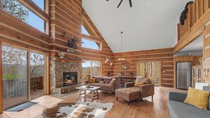 A spacious living area with a stone fireplace, wooden flooring, and a large window overlooking a deck.