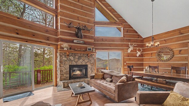 A log cabin interior with a stone fireplace, leather sofas, a dining table, and a view of the outdoors.