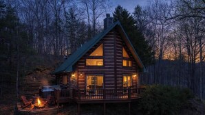 A log cabin with a deck and a fire pit, surrounded by a forest.
