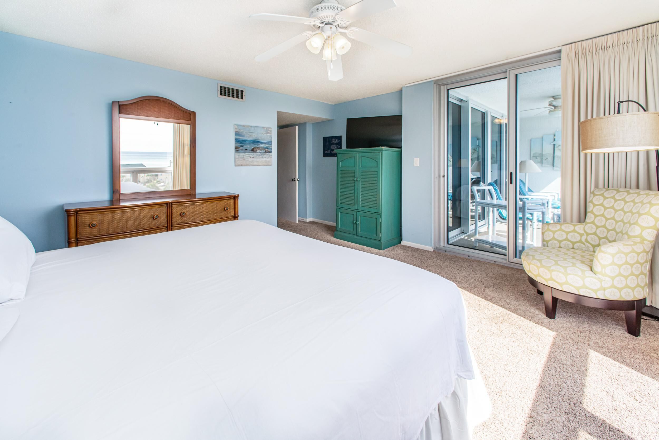 Une chambre avec un grand lit, une commode en bois, une armoire verte, une chaise et une vue sur l’extérieur, accessible par une porte-fenêtre coulissante.