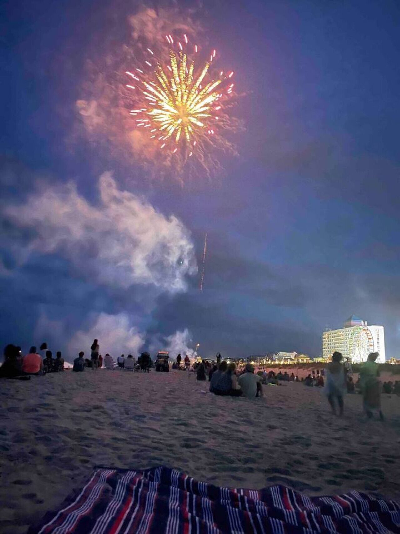 Ein Strand mit Menschen, die Feuerwerk beobachten. Im Hintergrund ist ein Gebäude mit einem Riesenrad zu sehen.