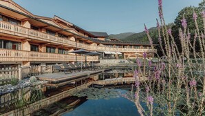 A multi-story wooden building with balconies, a clear water feature, and a mountainous backdrop. - Mariengut - Aparthotel Kramer (Wagrain)