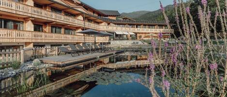 A multi-story wooden building with balconies, a clear water feature, and a mountainous backdrop.