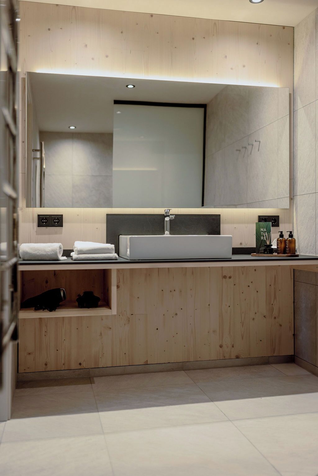 A modern bathroom with a large mirror, a white sink, and a wooden vanity.
