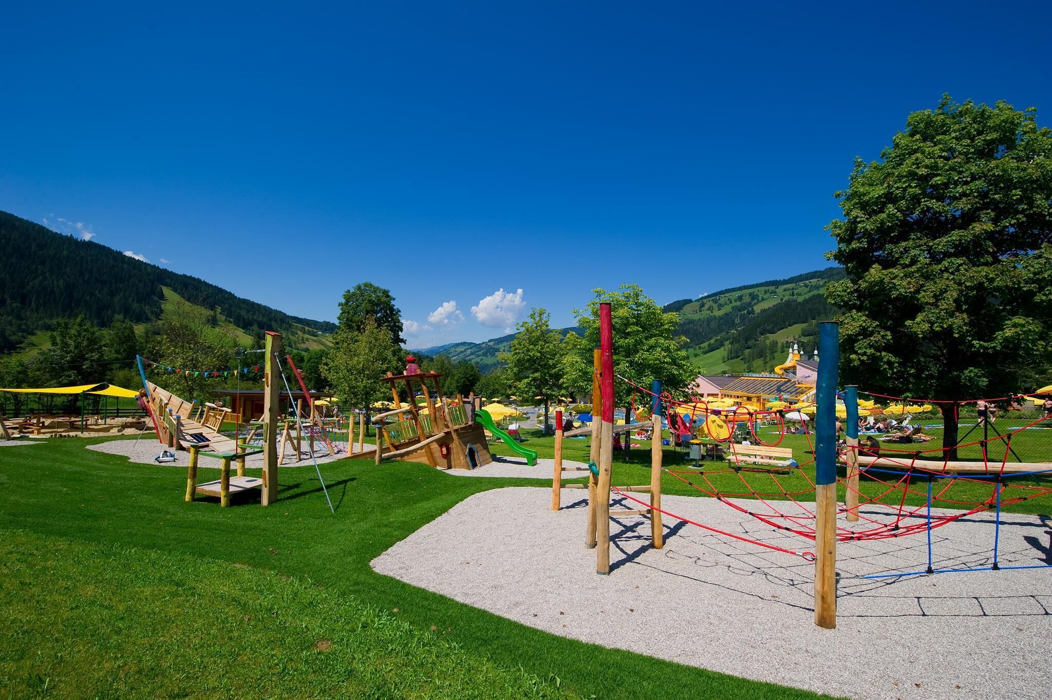 A playground with various play structures, a grassy area, and a mountainous backdrop.