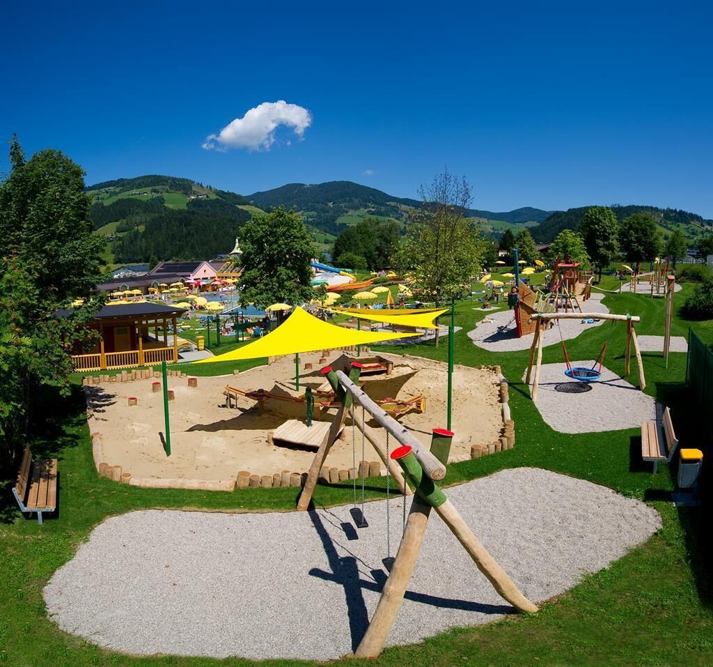 Un parque infantil con columpios, toboganes y un arenero, rodeado de césped y árboles.