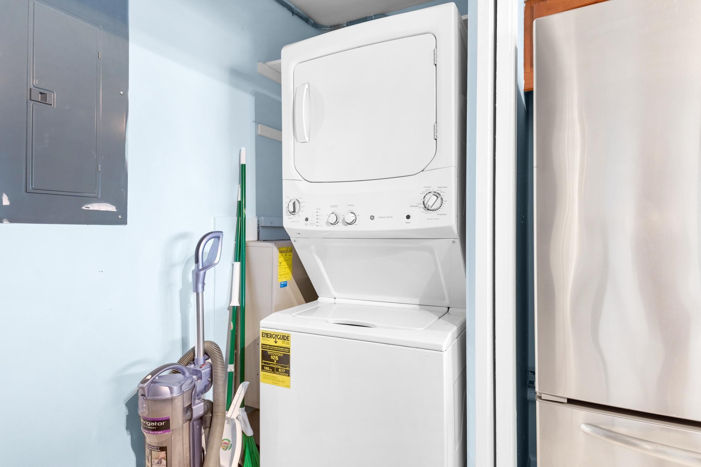 Une buanderie équipée d’un lave-linge et d’un sèche-linge superposés, d’un aspirateur et d’un réfrigérateur.