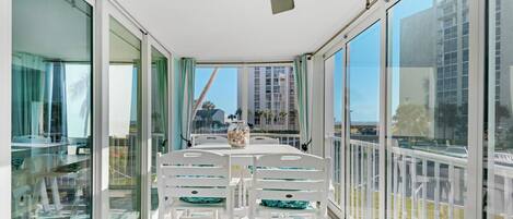 A sunlit balcony with a table and chairs, large windows, and a ceiling fan.