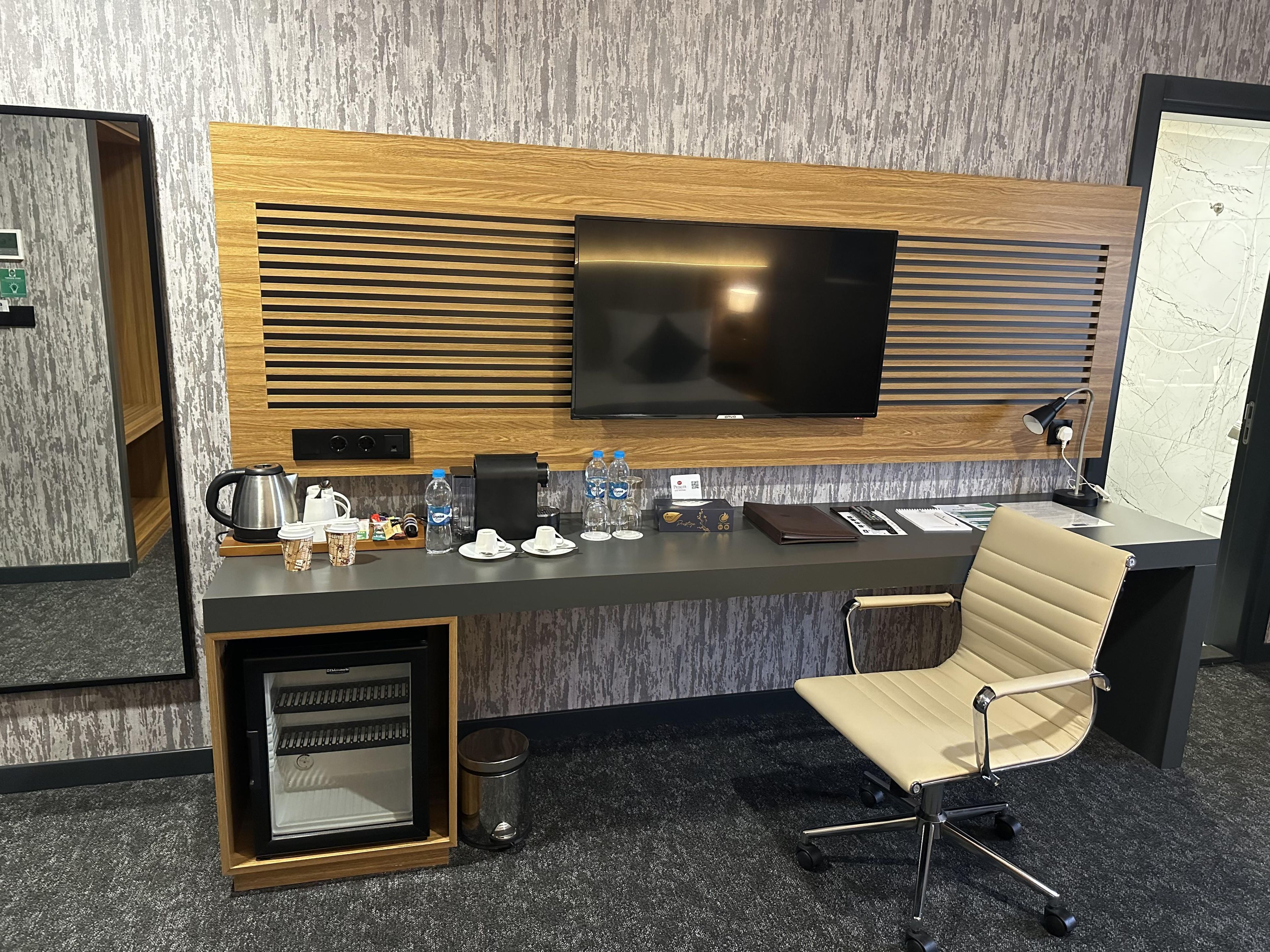 A modern hotel room with a flat-screen TV, a coffee station, a desk with a laptop, and a leather chair.