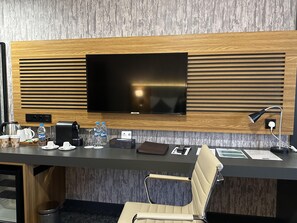 A modern hotel room with a large flat-screen TV mounted on a wooden panel wall, a desk with a chair, a coffee maker, a kettle, a water bottle, and a small table with a remote control and a phone. - Best Western Premier Dj Hotel (Djibouti)