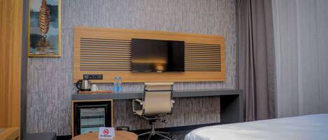 A hotel room with a bed, a desk with a television, a small table, a chair, a coffee maker, and a 'No Smoking' sign.