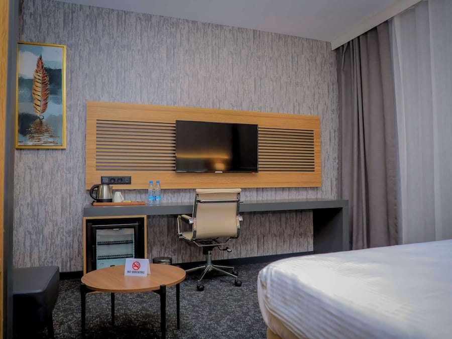 A hotel room with a bed, a desk with a television, a small table, a chair, a coffee maker, and a 'No Smoking' sign.