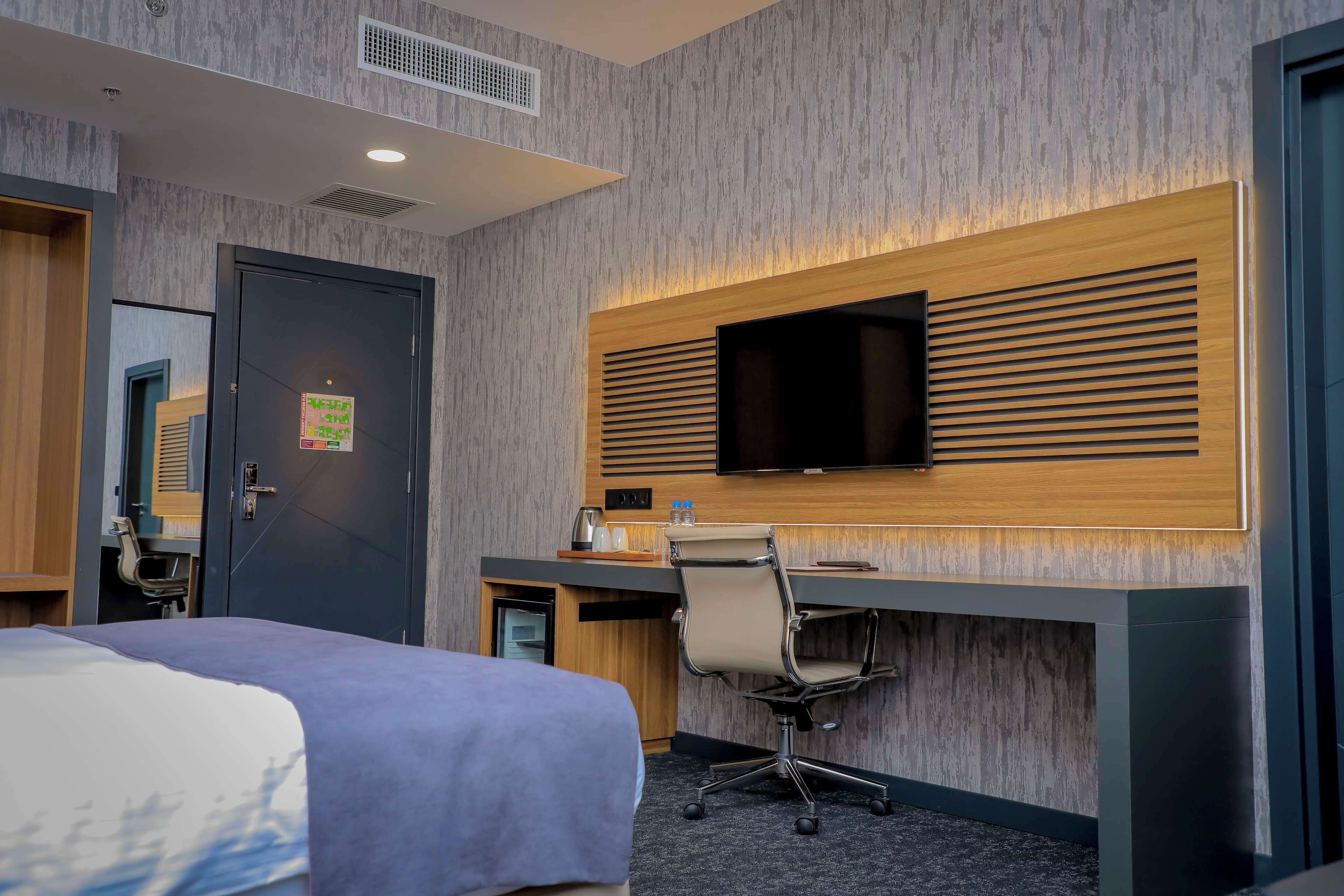 Habitación de hotel con cama, escritorio y silla, televisor montado en la pared y una puerta.