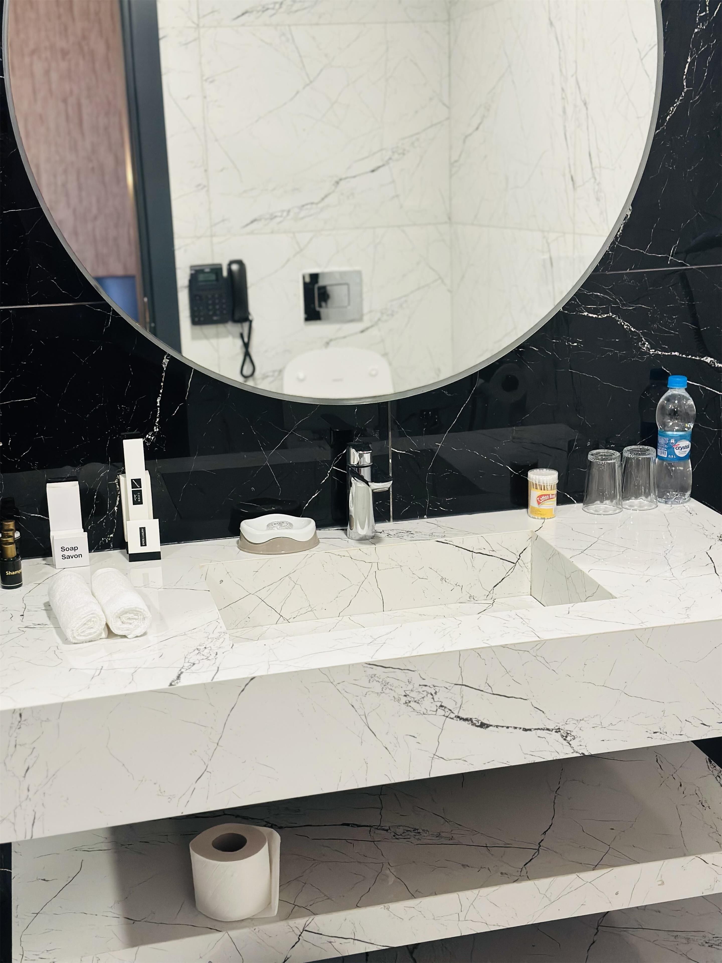 Un baño con encimera de mármol, espejo redondo, inodoro y diversos artículos de aseo.