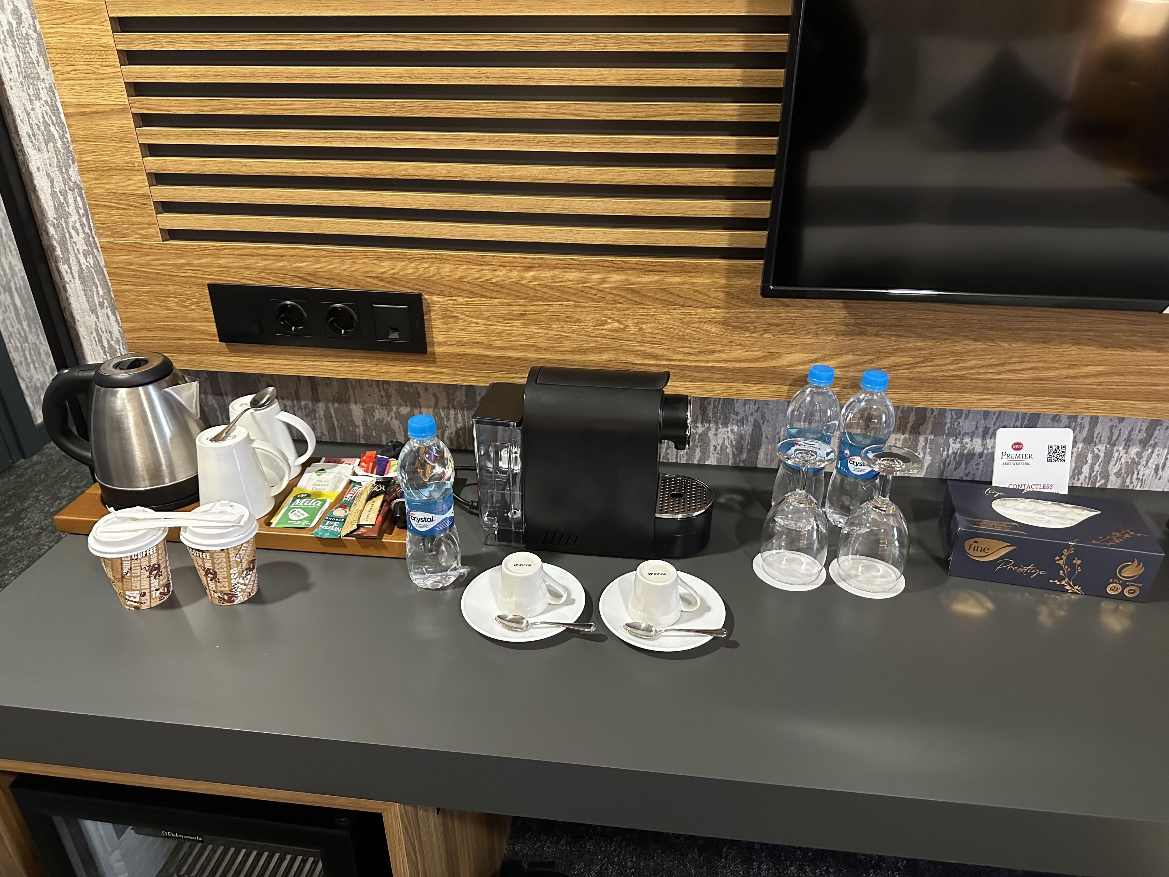 Una preparación para el desayuno con cafetera, hervidor de agua, botellas de agua y vasos desechables sobre una encimera.