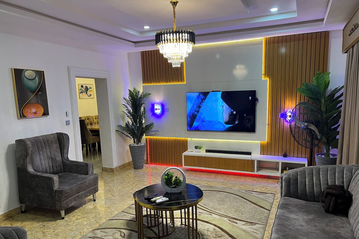 A modern living room with a large flat-screen TV, comfortable seating, a chandelier, and decorative plants.