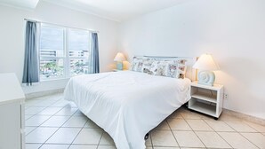 A hotel room with a large bed, two bedside lamps, a nightstand, a window with curtains, and a view of buildings outside. - Sea Oats 712 (Fort Walton Beach)