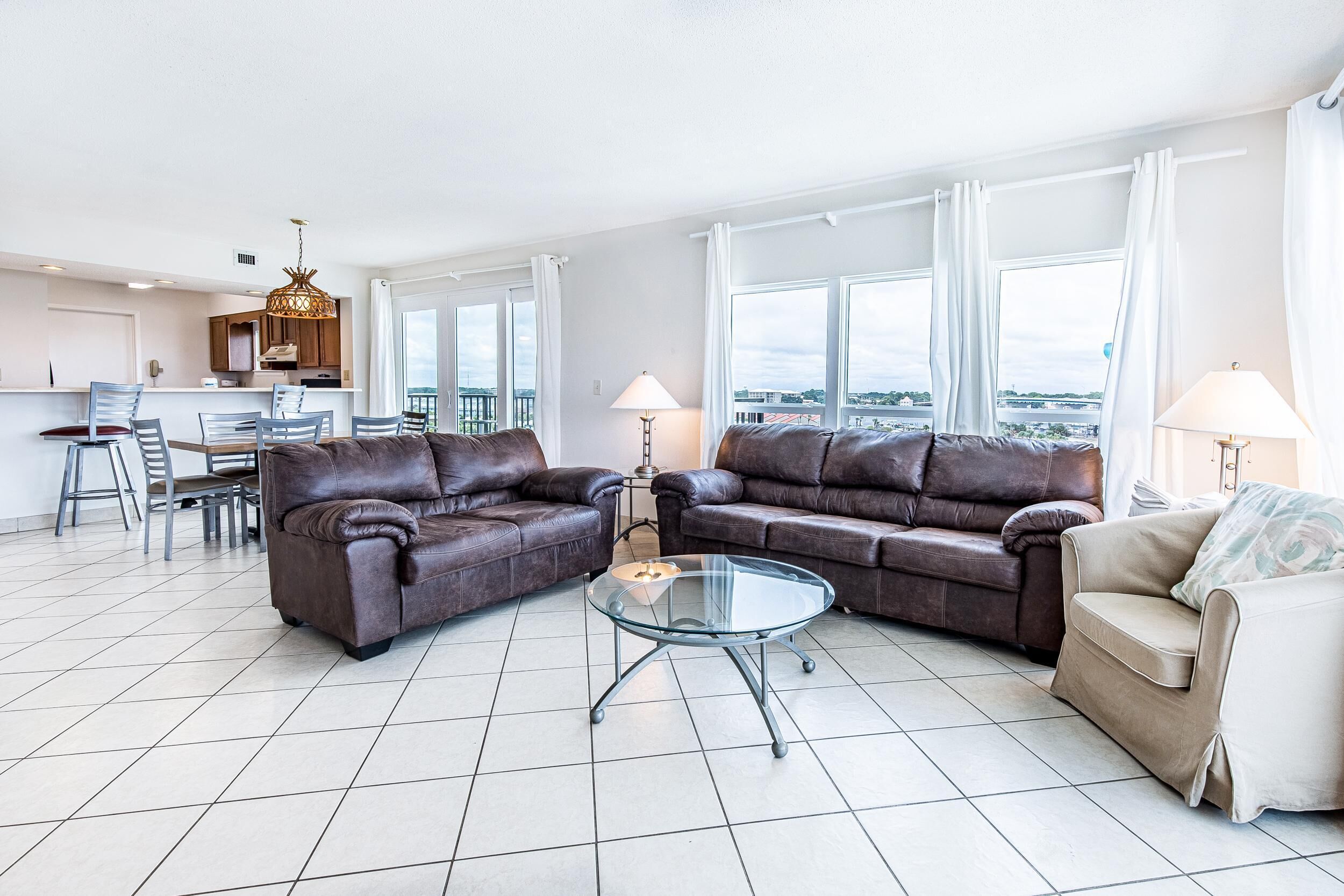 A modern living room with two brown leather sofas, a glass coffee table, and a beige sofa. There is a dining area with a table and chairs, and a kitchen in the background.