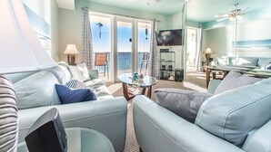 A spacious living room with a large sofa, a coffee table, and a television. There is a balcony with a view of the ocean.