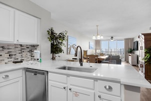 Cocina moderna con armarios blancos, lavavajillas y un lavabo. Hay un comedor con mesa y sillas, y vistas a la ciudad a través de grandes ventanales.