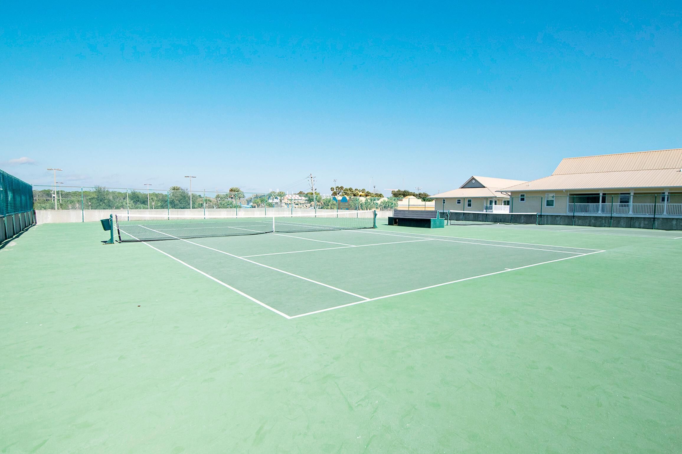 An outdoor tennis court with a net, surrounded by a low fence and a building with a flat roof.