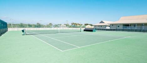 An outdoor tennis court with a net, surrounded by a low fence and a building with a flat roof.