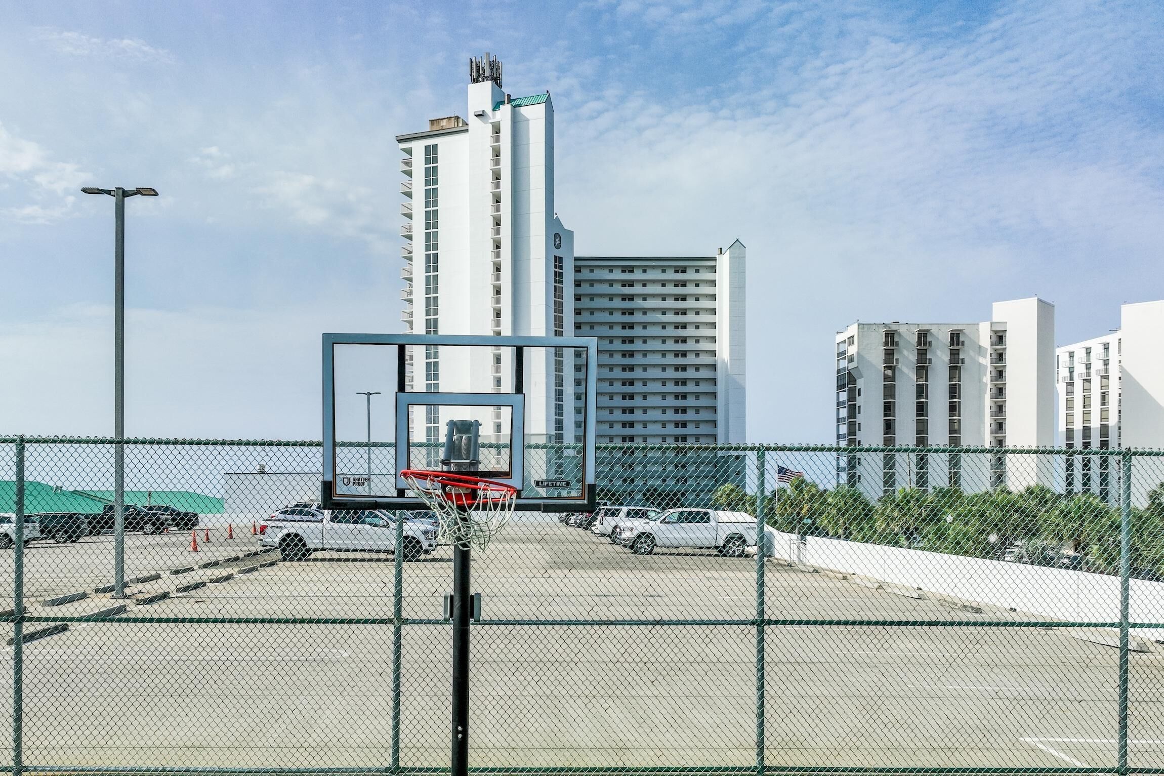 A basketball court with a chain-link fence, surrounded by parked cars and tall buildings in the background.