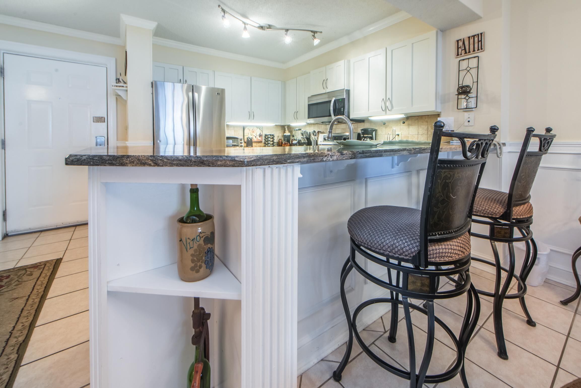 A kitchen with white cabinets, a granite countertop, and a stainless steel refrigerator. There are two bar stools with cushioned seats and black metal frames.