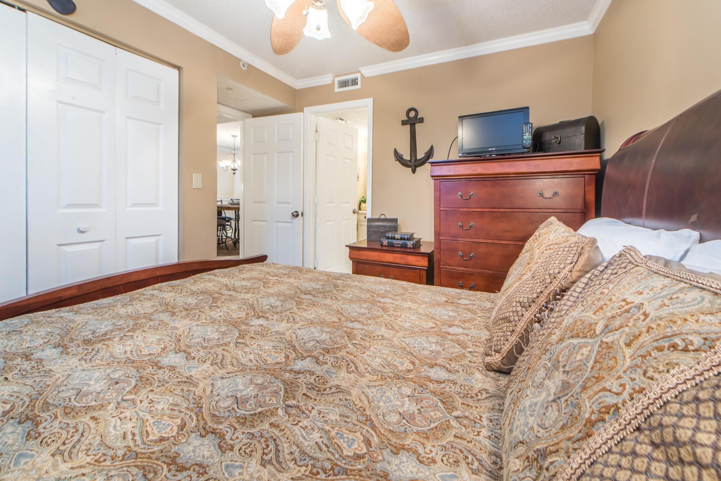 A bedroom with a patterned bedspread, a wooden dresser, a television, and an anchor decoration on the wall.