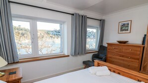 Hotellihuoneessa on suuri sänky, puinen lipasto, tuoli ja työpöytä, jossa on lamppu. Huoneessa on kaksi ikkunaa, joissa on verhot, ja seinällä on kehystetty taulu.