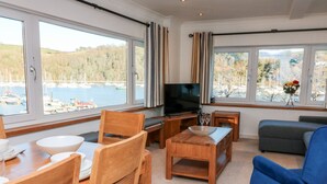 Huoneessa on puinen ruokapöytä, tuoleja, televisio, sohva ja suuret ikkunat, joista avautuu näkymä venesatamaan ja kukkuloille.