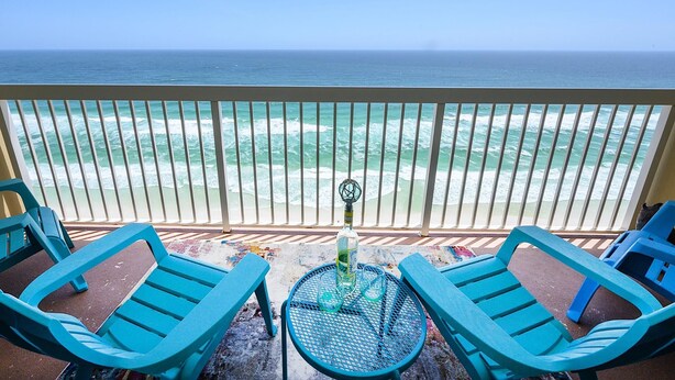 A balcony with two turquoise lounge chairs, a small round table, and a bottle of liquor. The view includes the ocean and a sandy beach.