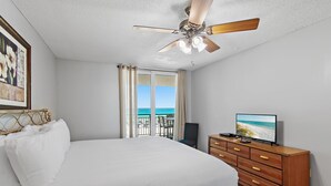 A bedroom with a bed, a ceiling fan, a television, a dresser, and a balcony with a view of the ocean.