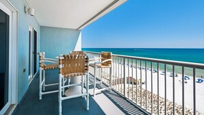 A balcony with two wicker chairs and a table, overlooking a beach with umbrellas and people. - Islander Beach Resort 6012 (Fort Walton Beach)