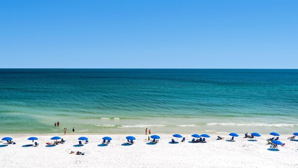 A beach with white sand and clear blue water. There are blue umbrellas and lounge chairs along the shore. - Islander Beach Resort 6012 (Fort Walton Beach)