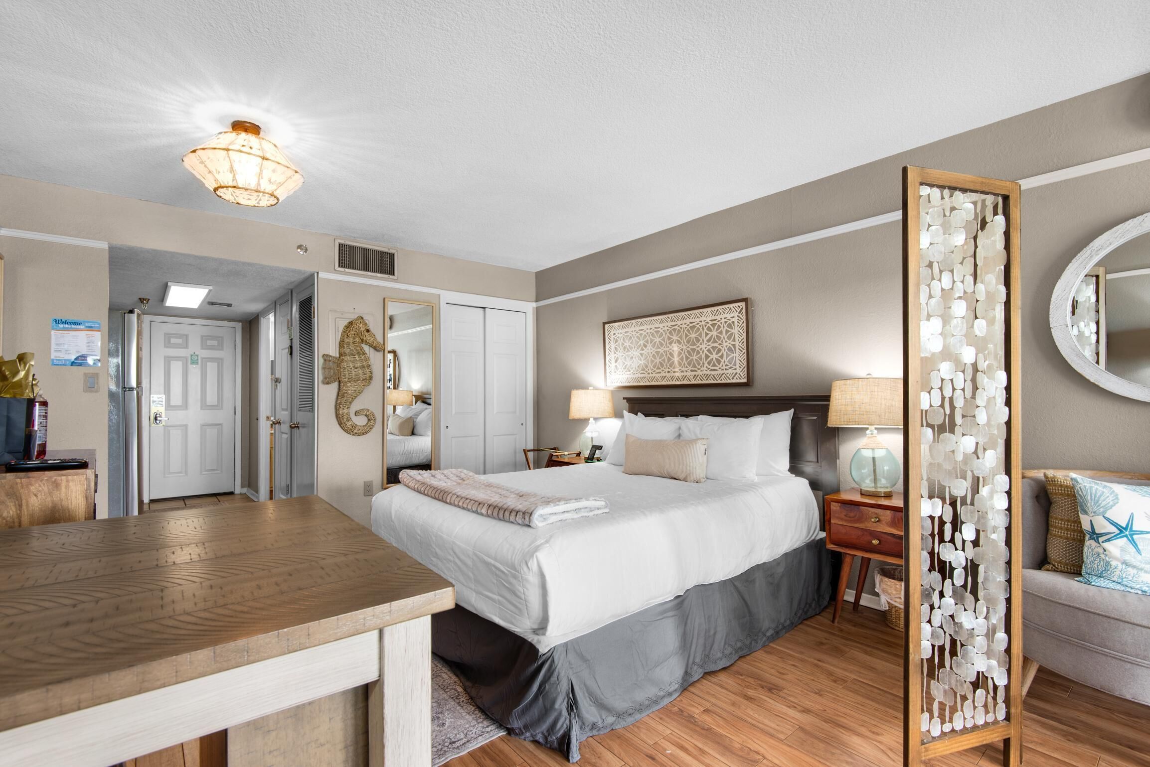 A hotel room with a bed, bedside table, lamp, decorative mirror, and a view of the hallway.