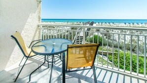 A balcony with a glass table and chairs, overlooking a beach with umbrellas and a clear blue sky.