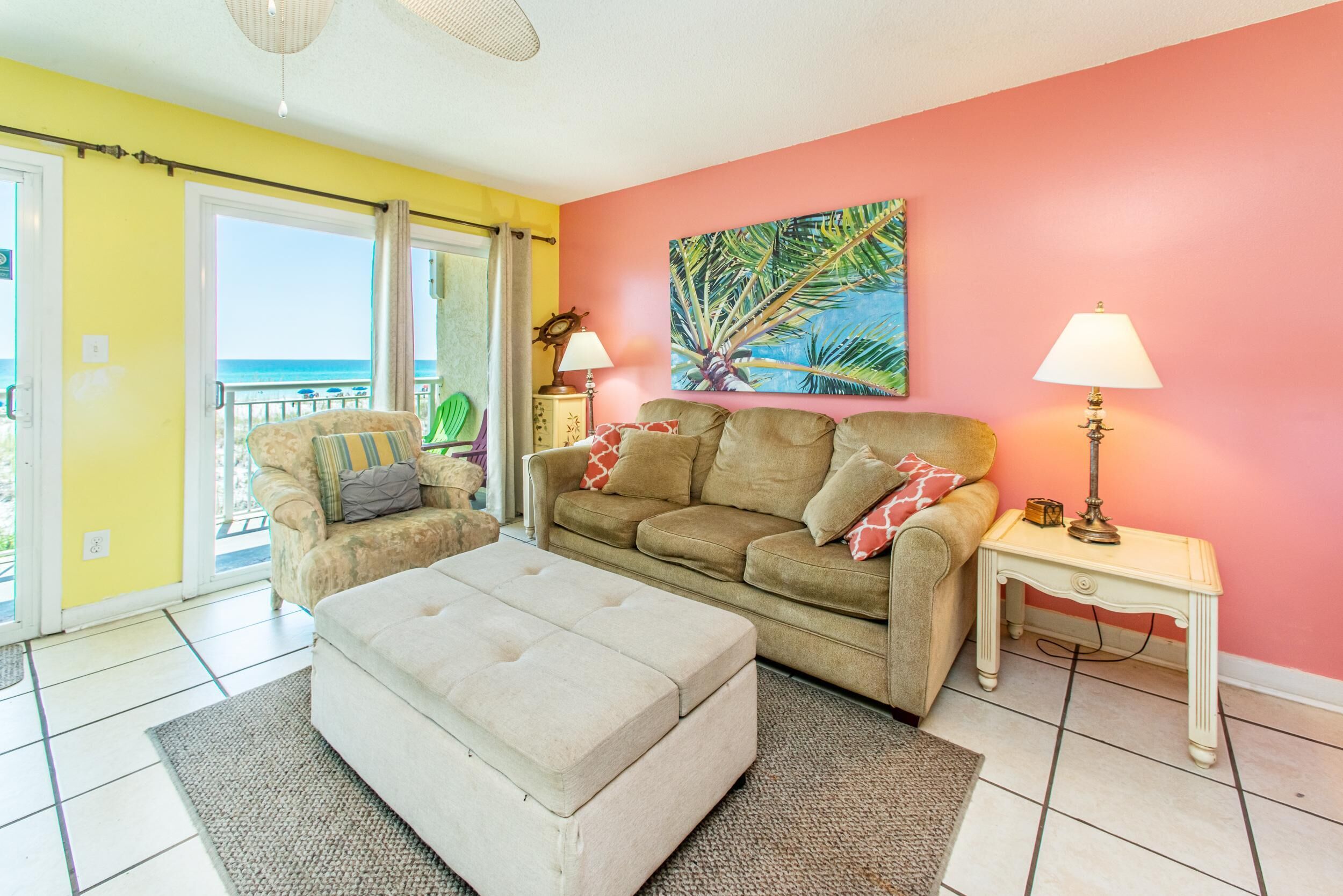 A living room with a sofa, ottoman, and a painting of a palm tree.