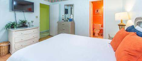 Un dormitorio con cama, cómoda, televisor y un baño visible a través de una puerta abierta.