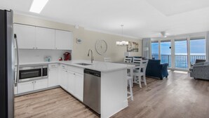 Et moderne køkken med hvide skabe, indbygget mikroovn og opvaskemaskine. Der er et spiseområde med et hvidt bord og stole, samt en balkon med havudsigt.