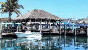 Een vastgelegde boot genaamd 'Sea Hunt' in een jachthaven met rieten dakgebouwen en blauwe parasols.