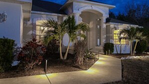 Een woonhuis met een overdekte patio, palmbomen en een goed verlichte oprit.