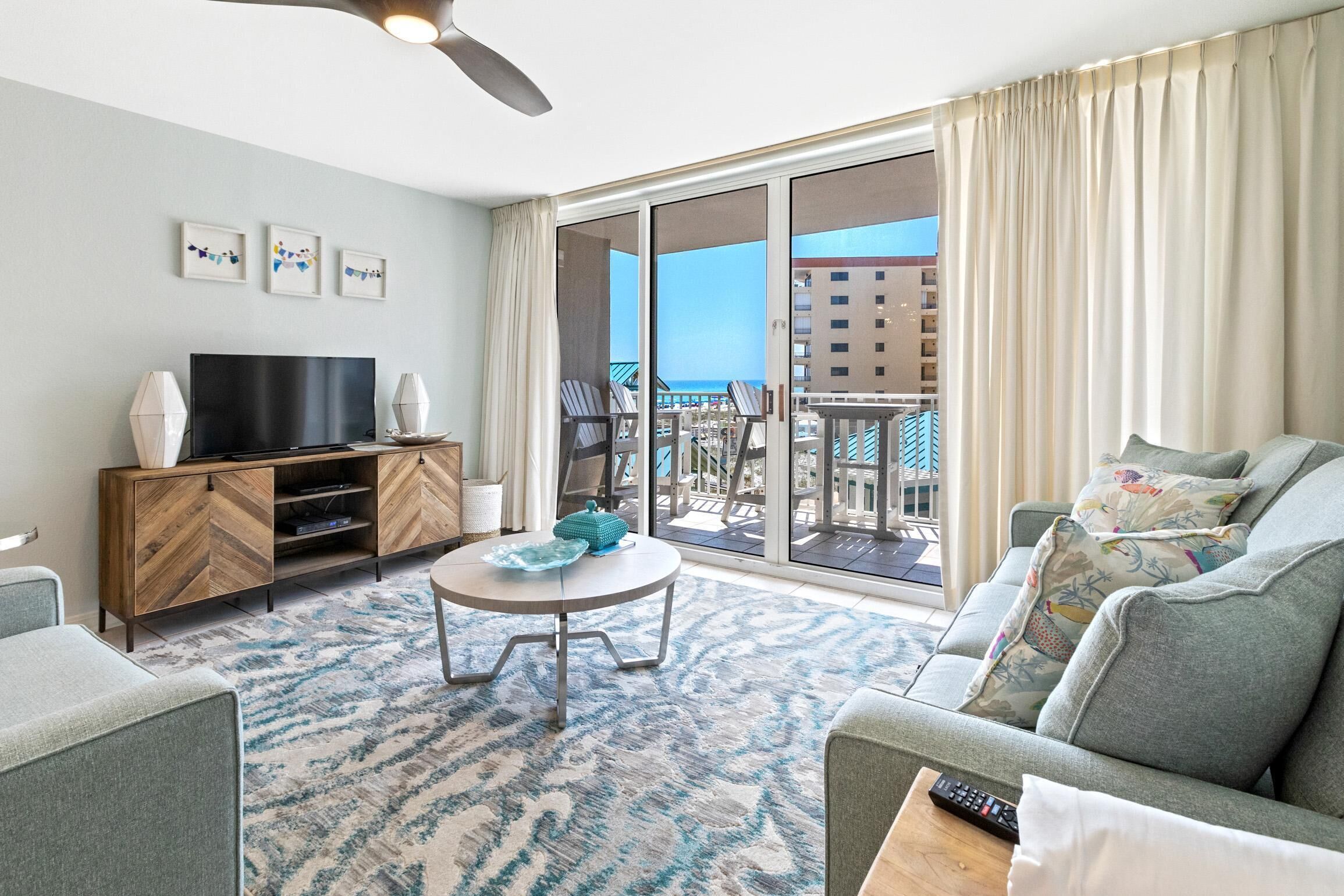 A modern living room with a flat-screen TV, a coffee table, and a sofa. There is a balcony with a view of the city and a pool.