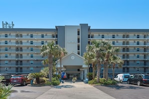 A multi-story hotel with a central entrance, surrounded by palm trees and parked cars.