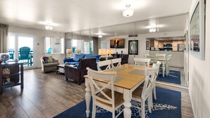 Amplia sala de estar con una mesa de comedor de madera, sillas blancas, un sofá azul y una cocina al fondo.
