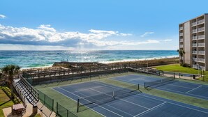 Ein Tennisplatz mit Blick auf den Ozean und den Strand, umgeben von einem Zaun und einem Gebäude.