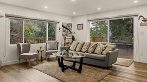 Una sala moderna con un sofá gris, dos sillones, una mesa de centro de vidrio y grandes ventanales que ofrecen vistas a una terraza y follaje.