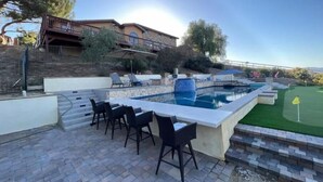 A pool area with a bar, lounge chairs, and a view of a house. - Meadowview Suite (Temecula)