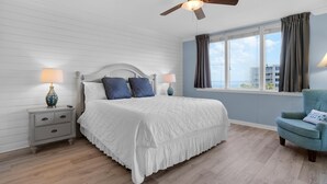Ein Schlafzimmer mit Bett, Nachttischen, einem Sessel, einem Deckenventilator und einem Fenster mit Vorhängen.