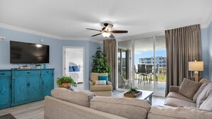 Ein Wohnzimmer mit Deckenventilator, einem Flachbildfernseher, einem blauen Schrank, einem beige farbenen Sofa, einem Couchtisch und Blick auf den Außenbalkon.